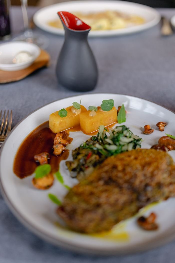 A gourmet dish featuring a main meat portion, potatoes, greens, and a dark sauce, elegantly plated.